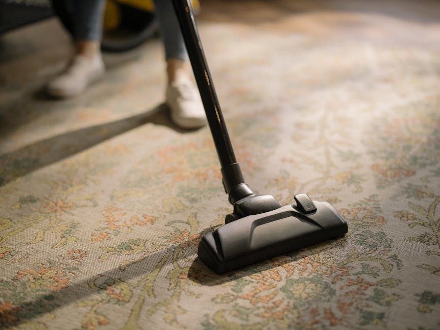 A person using a vacuum cleaner to clean a patterned, beige and green area rug in a well-lit room. The vacuum cleaner has a black body and a long, black handle, with visible controls near the base. Th