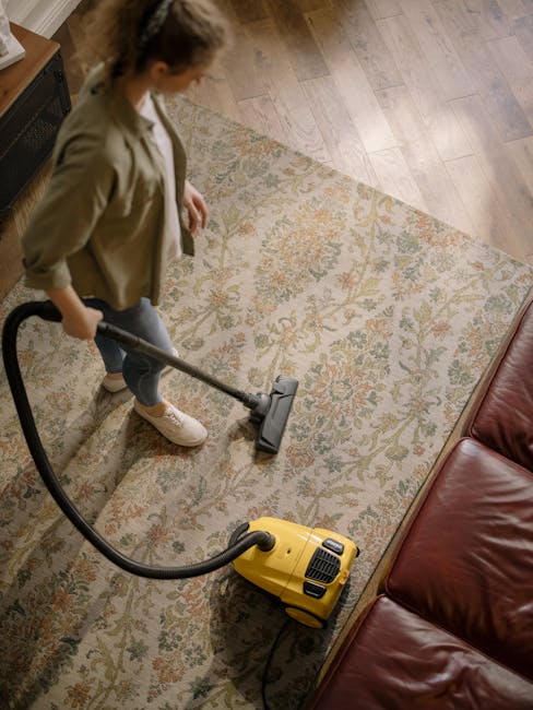 A person using a vacuum cleaner to clean a patterned, beige and green area rug in a well-lit room. The vacuum cleaner has a black body and a long, black handle, with visible controls near the base. Th