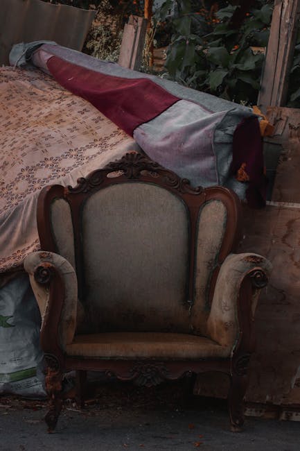 An antique wooden armchair with carved details and upholstered in light beige fabric, positioned in front of a wooden table draped with a patterned fabric. A rolled-up carpet or fabric material with shades of maroon, gray, and blue is placed on the table. The setting appears to be a rustic indoor space with wooden flooring and surrounding greenery outside a window or door, suggesting a mix of vintage furniture and household items. The area is well-lit with natural lighting, and the scene reflects a cleaning or decluttering environment that Carpet Cleaners Merton may address for surface cleaning and deep cleaning services.