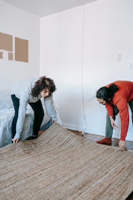 Two women are engaged in domestic cleaning activities in a well-lit room with white walls and a wooden floor. One woman, with curly hair and wearing a grey top and black pants, is lifting a large patterned area rug. The second woman, with dark hair tied back and wearing a red cardigan and grey pants, is seen adjusting the rug on the floor. Visible in the room are a white door, a power outlet, and a light switch on the wall. The scene depicts the process of surface cleaning or preparing to deep clean the carpet, supported by the presence of cleaning attire and the action of handling the rug, consistent with professional or thorough domestic carpet cleaning practices. Carpet Cleaners Merton provides expert cleaning services, including detailed carpet disposal and maintenance, ensuring hygienic and pristine surfaces.