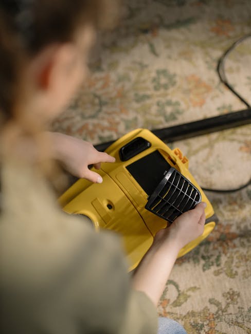 Close-up of a person using a black upright vacuum cleaner on a patterned, carpeted floor in a room with warm lighting. The vacuum cleaner's head is flat against the carpet, which features an intricate