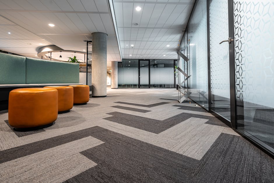 A spacious office lobby featuring patterned grey and beige carpet tiles with a geometric design, reflecting well-maintained cleanliness and hygiene standards. To the left, there are three rounded, orange leather ottomans placed near a sitting area with a green upholstered bench, adjacent to a concrete support column. The ceiling has built-in lighting and a curved design element, creating an inviting atmosphere. On the right, a glass wall with a hexagonal pattern partially reveals a room behind, and a metallic ladder is positioned against the glass. The overall scene emphasizes a modern, tidy environment suitable for professional business settings. This image illustrates the importance of surface cleaning and maintenance carried out by services like Carpet Cleaners Merton.