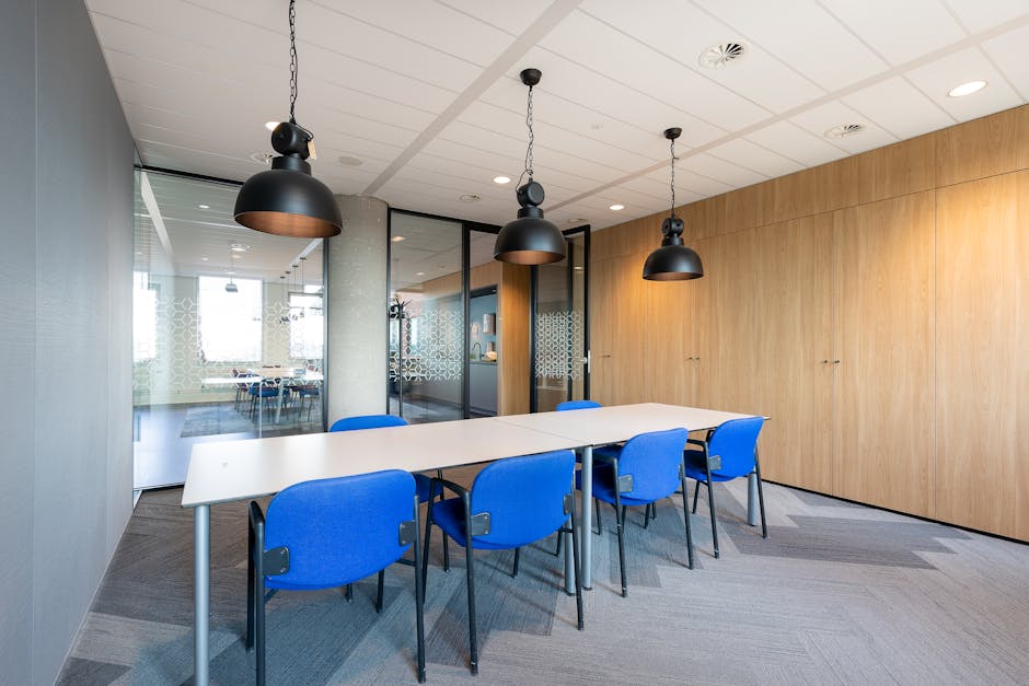 A modern conference room with a rectangular white table surrounded by six bright blue chairs featuring padded seats and armrests, situated on a grey carpeted floor. The room has a wooden wall panel on one side and a glass wall with decorative frosted patterns on the other, allowing natural light to illuminate the space. Three black pendant lights hang from the white ceiling with recessed lighting, creating a clean and professional atmosphere. The room appears neat and well-maintained, reflecting thorough surface cleaning and surface sanitisation carried out by Carpet Cleaners Merton as part of their commercial cleaning services for local businesses.