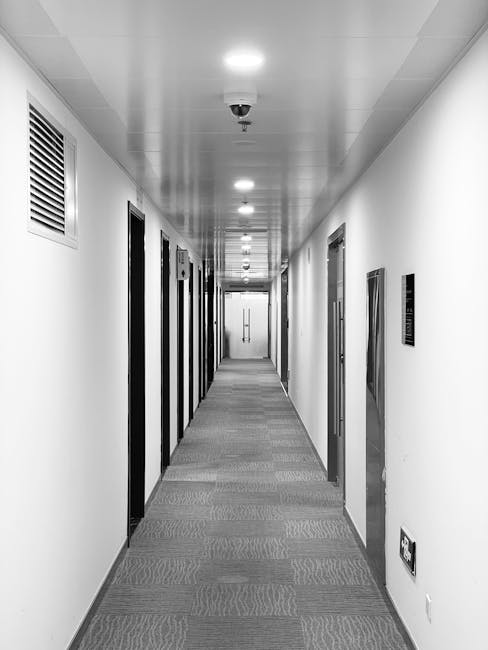 A spacious office lobby featuring patterned grey and beige carpet tiles with a geometric design, reflecting well-maintained cleanliness and hygiene standards. To the left, there are three rounded, orange leather ottomans placed near a sitting area with a green upholstered bench, adjacent to a concrete support column. The ceiling has built-in lighting and a curved design element, creating an inviting atmosphere. On the right, a glass wall with a hexagonal pattern partially reveals a room behind, and a metallic ladder is positioned against the glass. The overall scene emphasizes a modern, tidy environment suitable for professional business settings. This image illustrates the importance of surface cleaning and maintenance carried out by services like Carpet Cleaners Merton.
