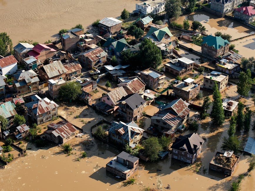 An aerial view of a residential area affected by flooding, showing many rooftops, some with rusted metal sheets, and streets submerged in muddy water. The area includes varied house styles with different roof materials, trees, and partial vegetation visible amidst the floodwaters, which have risen above the ground level, indicating significant flood impact. The scene is lit by daylight, highlighting the contrast between dry rooftops and flooded streets, with some homes appearing to have sustained water damage. The image emphasizes the importance of surface cleaning and sanitisation in flood-affected properties, as performed by companies like Carpet Cleaners Merton, to restore hygiene and property condition.