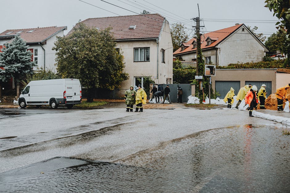 A residential street in Merton undergoing emergency flood response shows a wet, water-logged road surface with visible puddles and muddy patches. Several workers dressed in yellow and orange waterproof suits and helmets are engaged in surface cleaning and debris removal, using equipment and sandbags to manage flooding. In the background, there are detached houses with tiled roofs, some with windows, and a white van parked on the side of the street. Utility poles and overhead power lines are also visible, alongside trees and bushes. The scene depicts a coordinated effort to address flood damage, emphasizing thorough surface cleaning and sanitisation as part of emergency flood restoration services provided by Carpet Cleaners Merton.