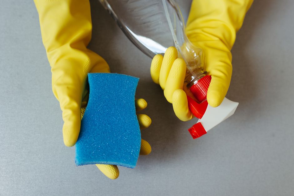 A variety of colorful cleaning product bottles and containers are arranged on a purple surface against a plain light grey wall, including spray bottles, squeezable bottles, and larger jugs in shades o