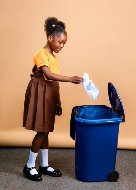 A young girl with dark skin, curly hair tied back, is standing indoors against a beige background. She is wearing a yellow short-sleeved top, a brown pleated skirt, white socks, and black shoes. The girl is holding a crumpled plastic bag over a blue wheeled trash bin, appearing to be in the process of discarding waste. The surface of the trash bin is smooth and shiny, and it is positioned on a grey carpeted floor. The scene is well-lit, emphasizing cleanliness and hygiene, which relates to proper waste disposal and surface cleanliness. This image exemplifies domestic or community waste management practices, aligning with the theme of responsible disposal under the guidance of Carpet Cleaners Merton for maintaining hygienic environments.