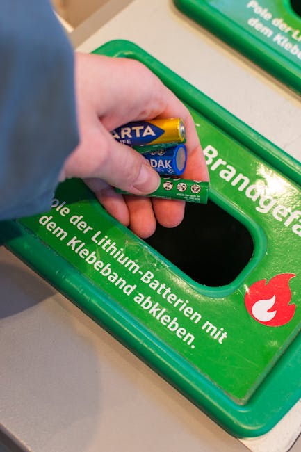Close-up of a person's hand inserting a large AA battery into a green recycling bin labeled with German text about lithium batteries disposal, situated on a white surface. The bin is part of a waste separation system, with a slot designed for battery recycling. The lighting is bright and even, highlighting the clean, glossy surface of the bin and the bright yellow and blue colors of the battery. The setting appears to be indoors, emphasizing environmental responsibility and proper waste management practices relevant to residential or commercial cleaning standards, as promoted by Carpet Cleaners Merton for maintaining hygienic spaces. The background is slightly blurred, focusing on the battery and recycling bin, illustrating proper disposal procedures in line with waste rules and emphasising hygiene and maintenance of clean environments.