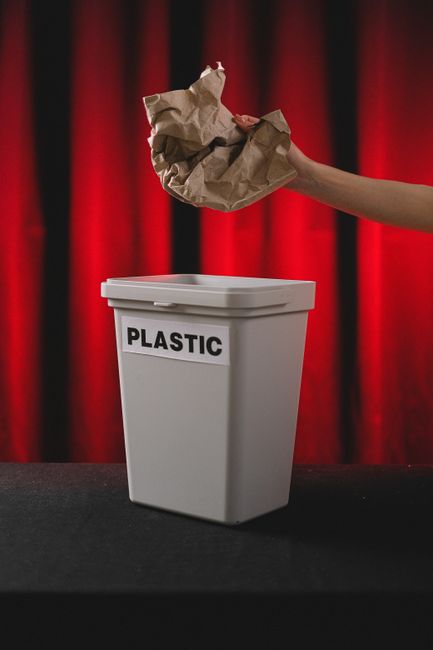 A young girl with dark skin, curly hair tied back, is standing indoors against a beige background. She is wearing a yellow short-sleeved top, a brown pleated skirt, white socks, and black shoes. The girl is holding a crumpled plastic bag over a blue wheeled trash bin, appearing to be in the process of discarding waste. The surface of the trash bin is smooth and shiny, and it is positioned on a grey carpeted floor. The scene is well-lit, emphasizing cleanliness and hygiene, which relates to proper waste disposal and surface cleanliness. This image exemplifies domestic or community waste management practices, aligning with the theme of responsible disposal under the guidance of Carpet Cleaners Merton for maintaining hygienic environments.