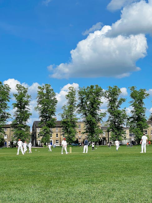 A group of people playing cricket on a well-maintained grassy field adjacent to a row of residential buildings, with tall green trees lining the background under a bright blue sky with fluffy white clouds. The scene depicts outdoor recreation and leisure activities, with players in white uniforms actively engaged in the game. The natural lighting highlights the lush grass and the clear sky, emphasizing a clean and inviting environment suitable for outdoor sports. This image showcases the importance of outdoor surface maintenance and cleanliness that Carpet Cleaners Merton can support through professional cleaning services, especially in residential or communal outdoor spaces.