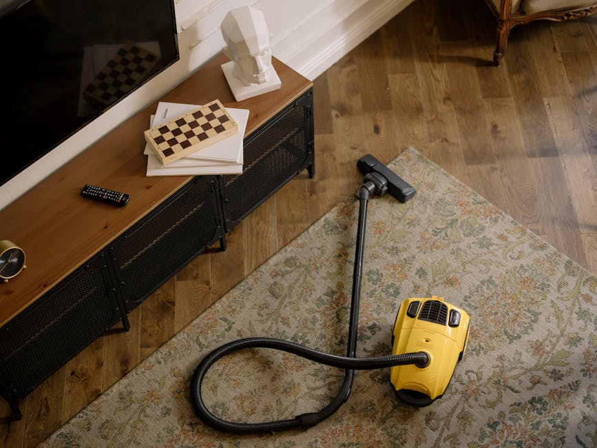 A professional cleaning technician dressed in full protective gear, including a white hazmat suit, face mask, gloves, and shoe covers, is performing surface cleaning on a beige, textured carpet in a modern, well-lit living room. The room features a large mirror leaning against a white wall, potted plants, and wooden shelving with decor items and books. Using a steam cleaning machine, the technician is applying deep cleaning to remove dirt and stains from the carpet, which appears moist with steam generated by the equipment. The scene emphasizes thorough sanitisation and maintenance of domestic carpets, showcasing expertise in deep cleaning services provided by Carpet Cleaners Merton near Cricket Green, Merton.
