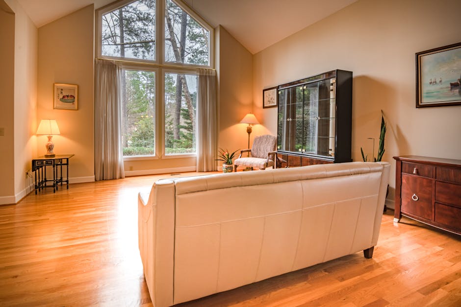 A spacious living room with light-colored wooden flooring and cream walls, illuminated by natural light from large, multi-pane windows dressed with white curtains. In the center, a white upholstered sofa faces away from the camera, with a small wooden side table holding a traditional lamp with a beige shade on the left. To the right, a wooden cabinet with a glossy finish and metal handles is positioned against the wall, and a framed landscape painting hangs above it. An armchair near the window is decorated with a potted plant. The room appears clean, tidy, and well-maintained, reflecting comprehensive surface cleaning and domestic cleaning practices by Carpet Cleaners Merton, as seen in this inviting, well-lit space.