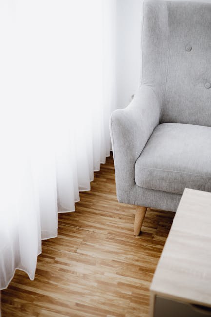 A corner of a residential room featuring a light grey upholstered armchair with wooden legs positioned next to sheer white curtains that hang to the floor. The room has a wooden floor with a natural finish, appearing clean and well-maintained. A small light-colored wooden table is partially visible in the foreground. The lighting is soft, likely natural daylight filtering through the curtains, highlighting the tidy and inviting atmosphere. This setting exemplifies domestic cleaning and surface maintenance, with emphasis on furniture and flooring hygiene. Carpet Cleaners Merton offers professional deep cleaning and upholstery sanitisation for homes in Morden, as suggested by the context of the Morden SM4 Guide page.