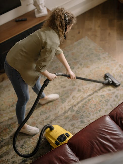 A young boy using a vacuum cleaner to deep clean a beige sofa with two cushions in a living room. An elderly man with gray hair, wearing a gray long-sleeve shirt and dark pants, is lying on the sofa, resting with his hand on his forehead. The room features large windows with sheer curtains, wooden flooring, and a bright yellow and gray rug. A black side table with a lamp is positioned beside the sofa, and a white radiator is visible beneath the windows. Visible cleaning equipment includes the vacuum cleaner being operated by the boy and an additional vacuum device on the floor. The scene showcases domestic cleaning with a focus on surface cleaning and hygiene maintenance, reflecting the services promoted by Carpet Cleaners Merton for high-quality deep cleaning and sanitisation near SW19, Wimbledon High Street.
